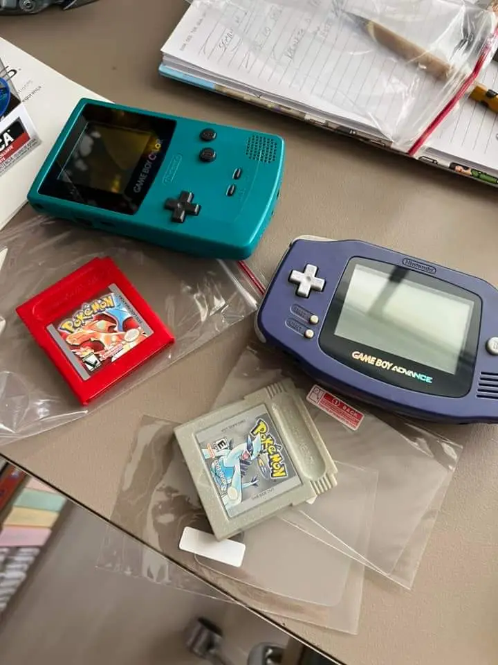 A teal Game Boy Color and a blue Game Boy Advance sit on a desk next to two Pokémon game cartridges in plastic sleeves, one…
