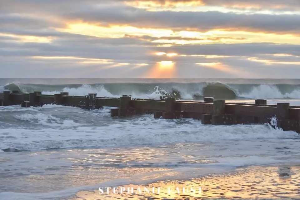 A scenic photograph of a stormy ocean at sunset, with waves crashing against a wooden breakwater. The sky is filled with…