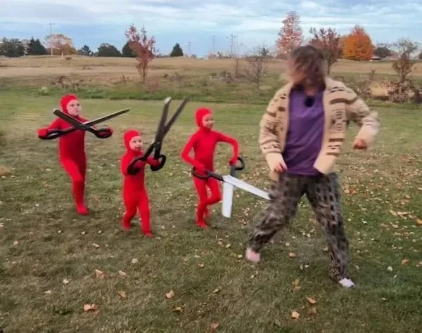 Four children in red full-body costumes holding oversized scissors stand in a grassy field facing a person in a beige jacket…