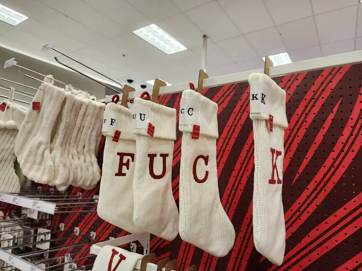 Four white knit Christmas stockings hang in a retail store, each displaying a large red letter spelling 'F', 'U', 'C', 'K'…