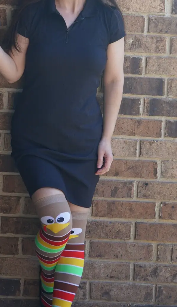 A person wearing a dark blue dress stands against a brick wall, showcasing colorful striped knee-high socks with a cartoon…