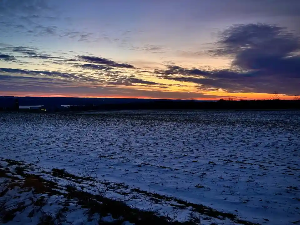 A snowy field at dusk with a vibrant orange and purple sunset sky, silhouetted farm buildings in the distance. The overlaid…