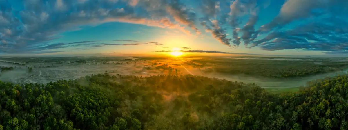 A panoramic aerial view of a sunrise over a misty landscape with forests, fields, and distant suburbs. The sun is low on the…