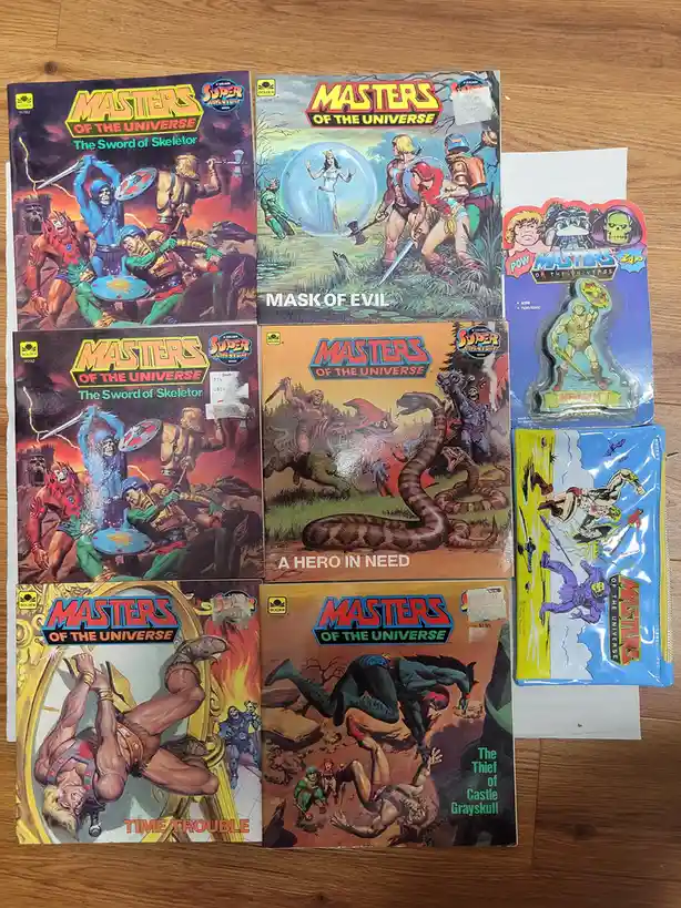 A collection of vintage 'Masters of the Universe' comic books and toys laid out on a wooden surface. The comics feature…