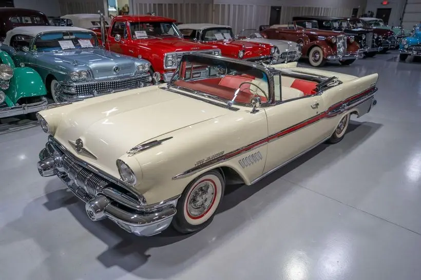A cream-colored 1950s convertible classic car with red interior and chrome details is prominently displayed in a showroom…