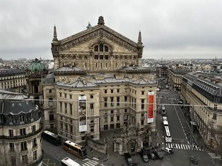 An aerial view of the Paris Opera House on an overcast day, showing its grand neoclassical facade, surrounding streets with…