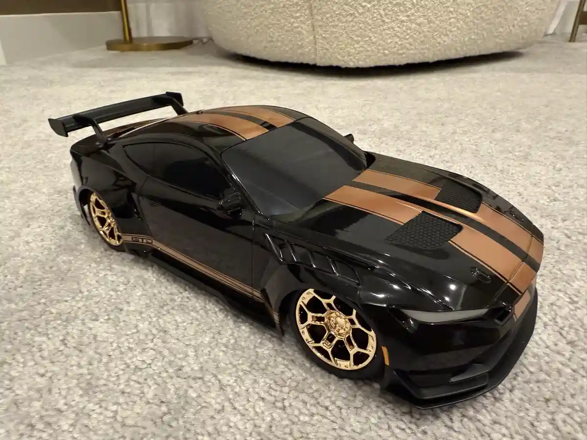 A black die-cast model of a Ford Mustang GT with gold and bronze racing stripes, large rear wing, and gold wheels sits on a…