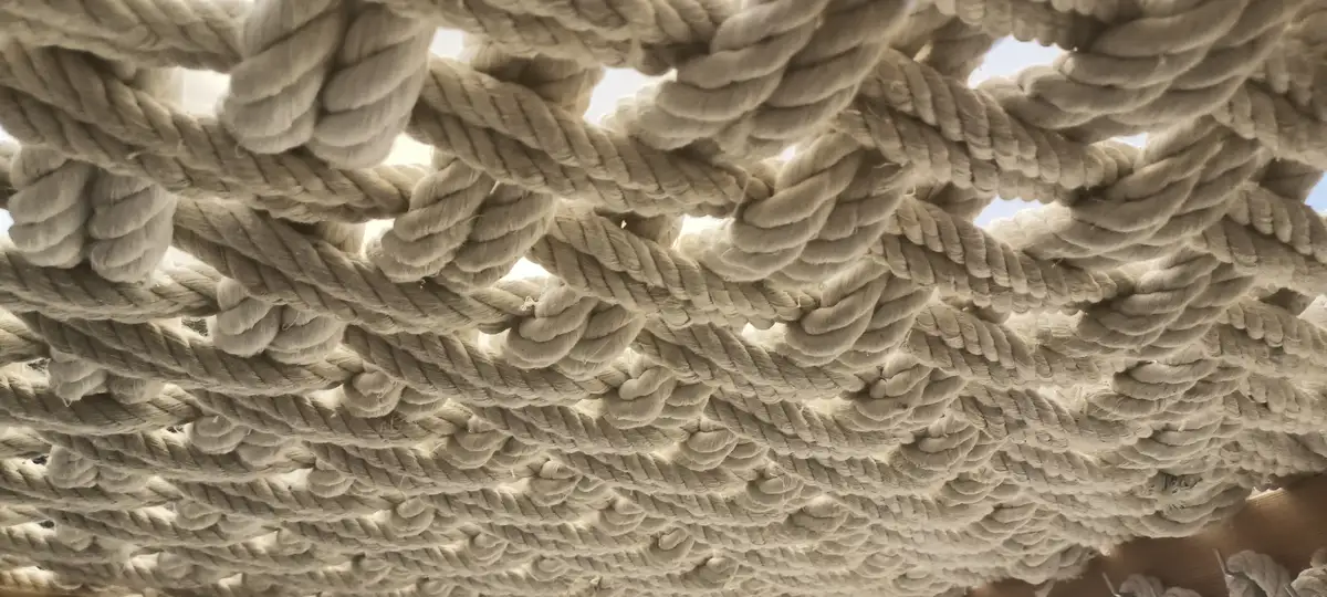 A close-up photo shows thick, beige ropes woven together to form a ceiling or overhead structure, with patches of bright sky…