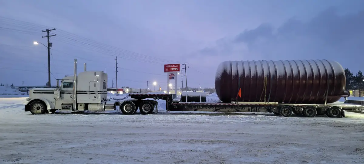 A white semi-truck with a flatbed trailer is parked in a snowy lot at dusk, hauling an enormous, ribbed, cylindrical tank…