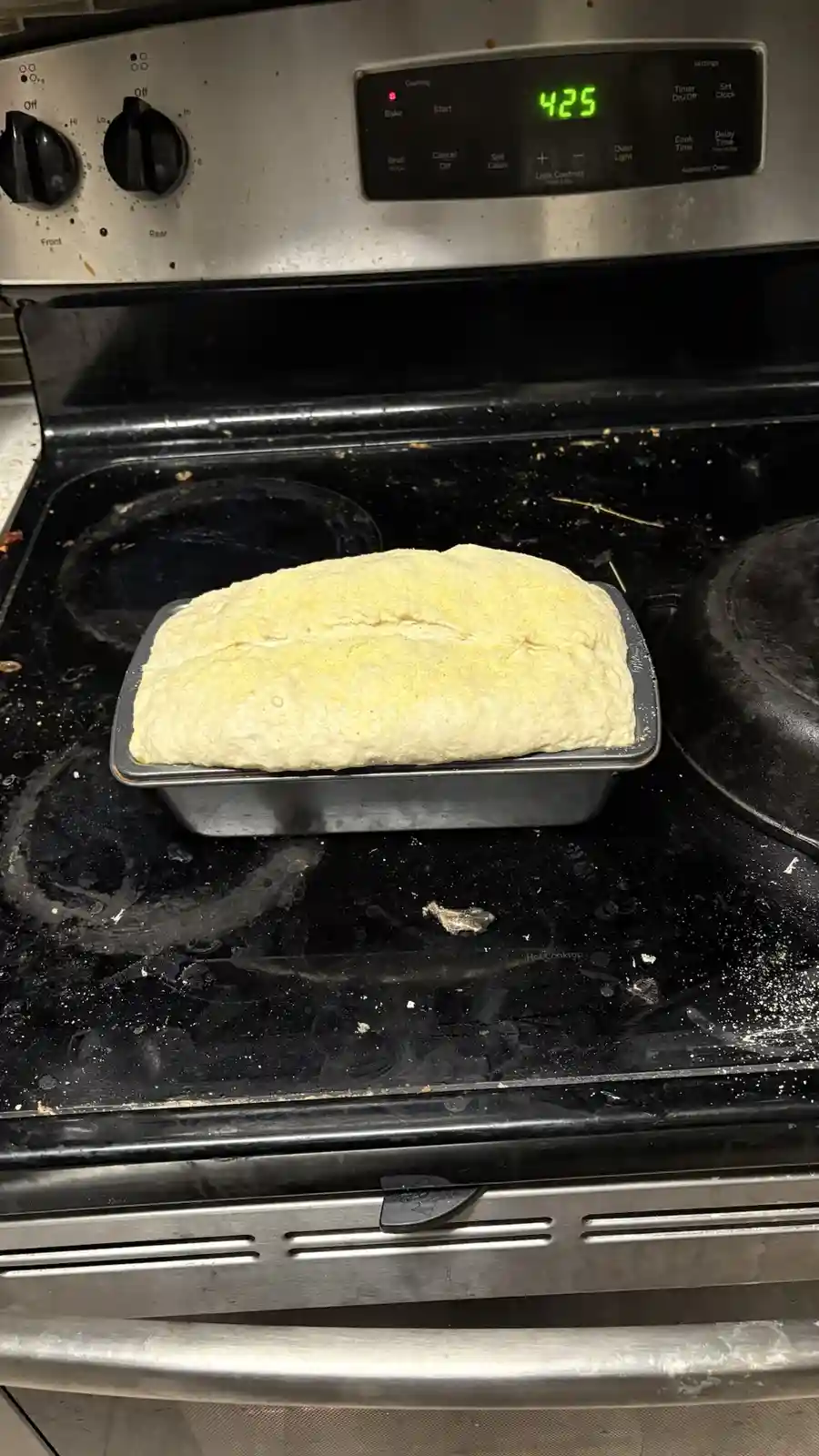 A loaf of unbaked bread dough sits in a metal pan on a black electric stovetop, with the oven's digital display clearly…