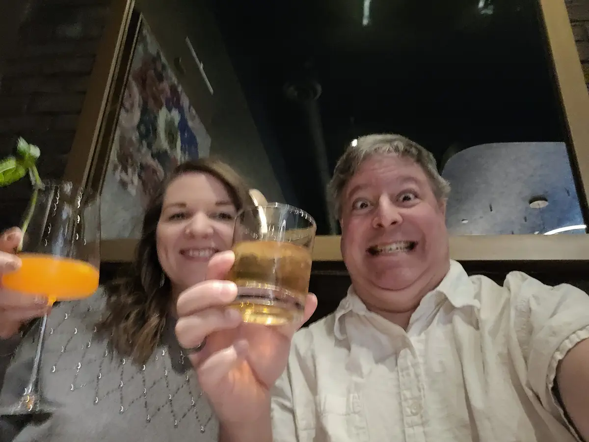 A man and woman are taking a selfie at a bar or restaurant, both holding up drinks. The woman holds a cocktail in a coupe…