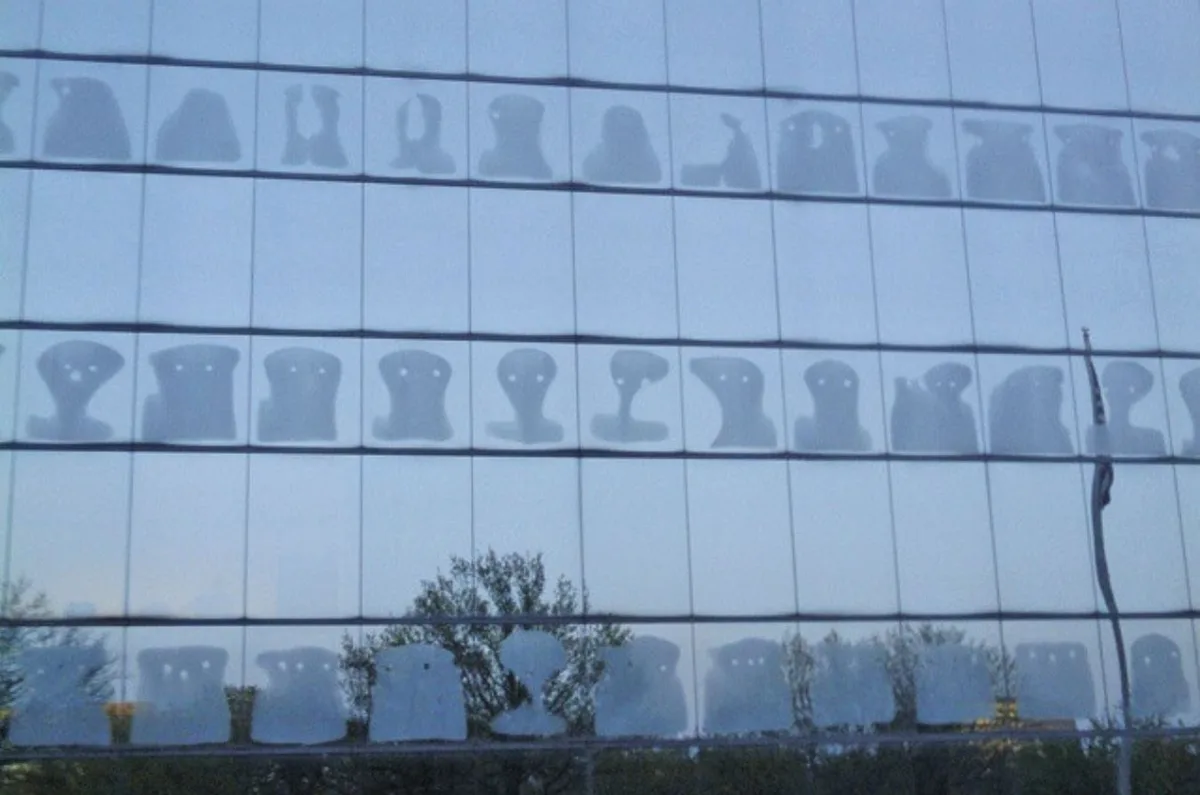 A glass office building facade reflects the silhouettes of many people sitting at desks, their heads and shoulders visible…
