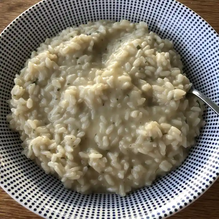 A close-up photo shows a bowl of creamy risotto with visible rice grains and green herbs, served in a white bowl with a blue…