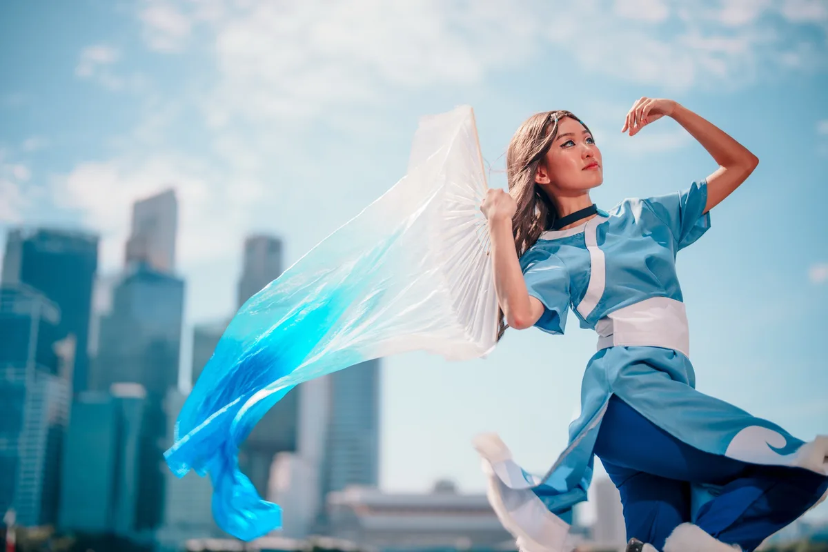 A person in a blue and white Water Tribe cosplay from Avatar: The Last Airbender poses dynamically against a city skyline…