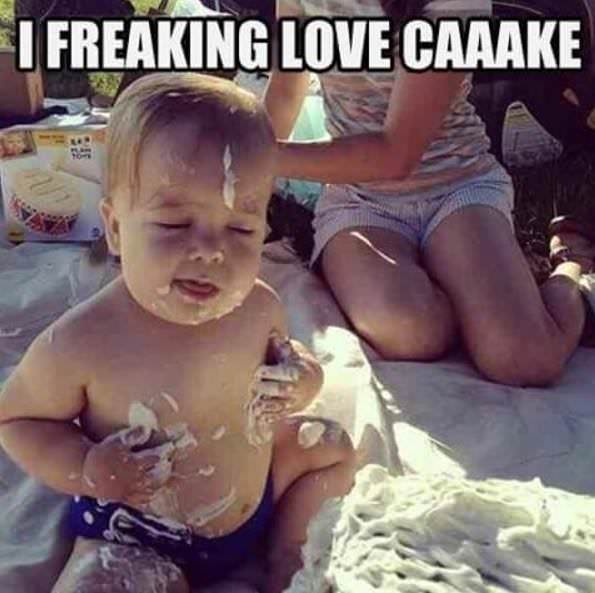 A baby with frosting smeared on their face and hands sits on a blanket, covered in cake, while an adult sits behind them…