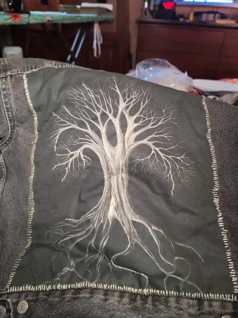 A close-up photo shows the back of a dark denim jacket with a large, intricate white embroidery of a tree that has visible…