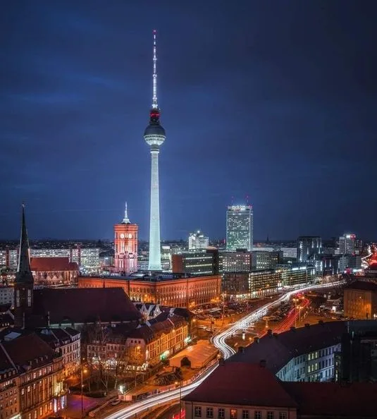 A nighttime cityscape photograph of Berlin, featuring the iconic Fernsehturm (TV Tower) illuminated against a dark blue sky…