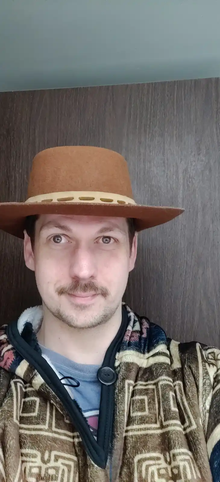 A man with a mustache wears a brown cowboy hat and a patterned fleece blanket hoodie while looking directly at the camera…