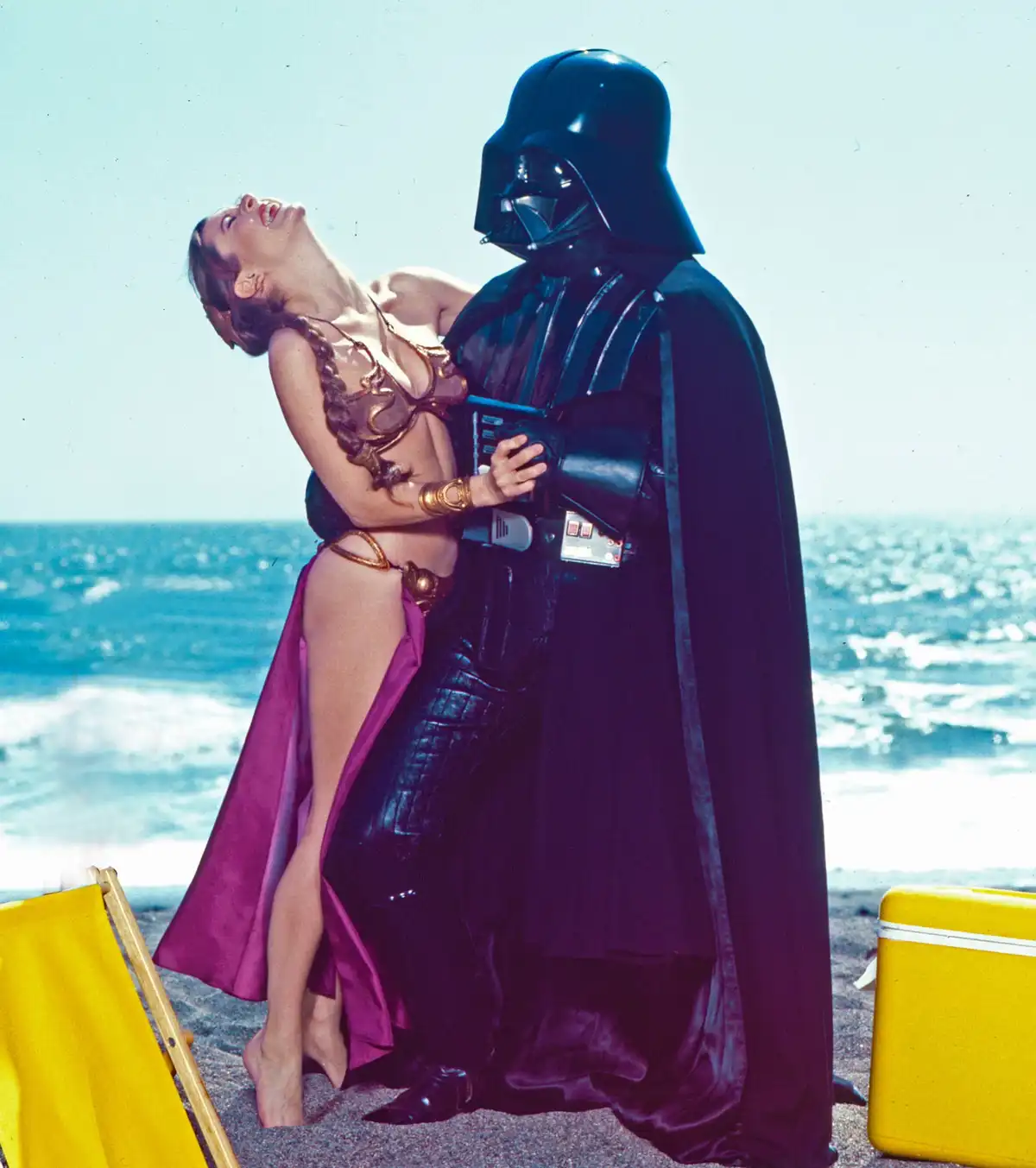 A still from Star Wars shows Darth Vader holding Princess Leia on a beach, with a yellow beach chair and cooler nearby. The…