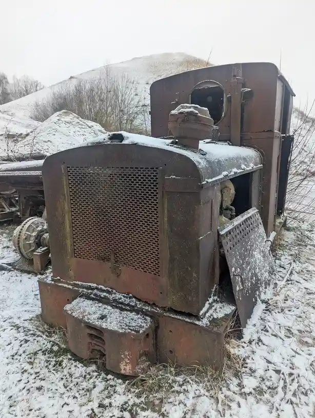 A rusted, snow-dusted old industrial engine sits abandoned in a snowy field with piles of snow-covered debris behind it. The…