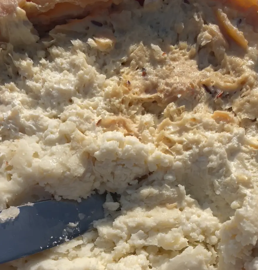 A close-up photo shows a lumpy, pale, textured food substance being scraped with a knife, resembling mashed potatoes or a…