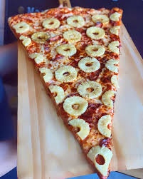 A large, triangular slice of pizza topped with numerous pineapple rings sits on a wooden serving board. The pizza has a…