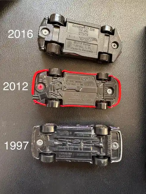 Three Hot Wheels toy cars are shown upside down on a textured black surface, each labeled with a year: 2016 (Corvette C7…