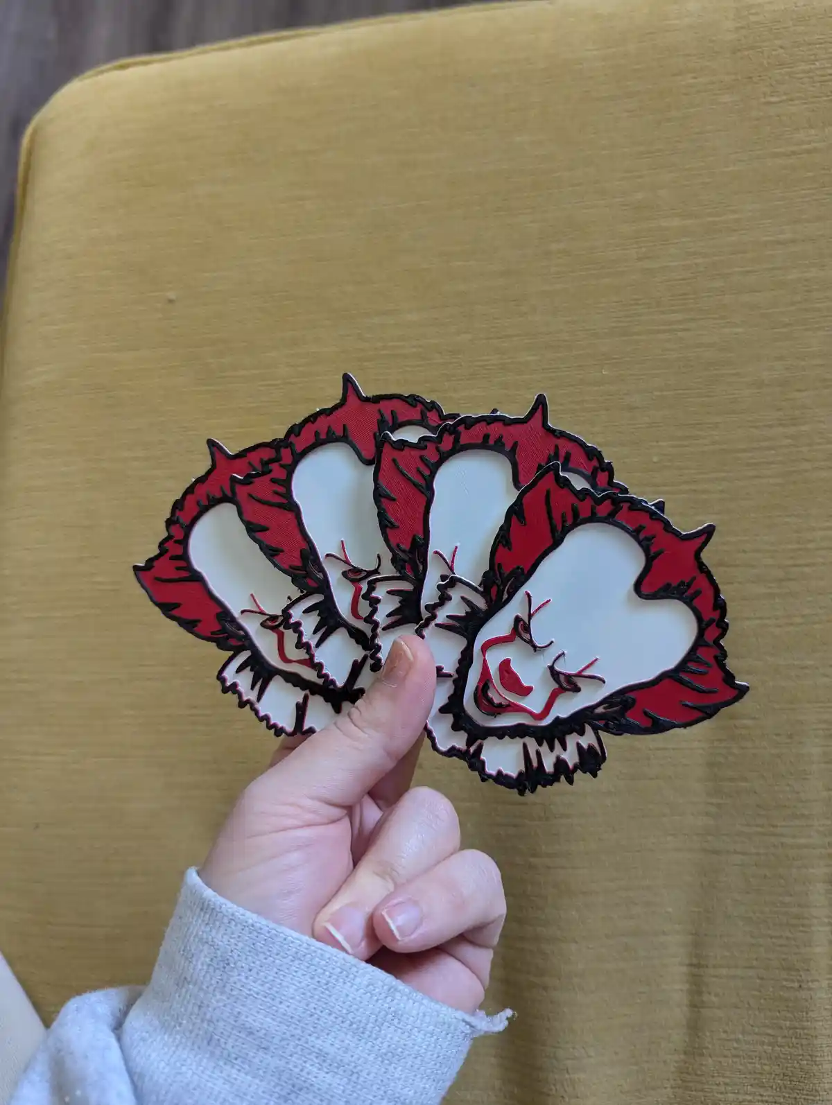 A hand holds three identical red-and-white 3D-printed fan-shaped cutouts, each depicting a stylized, angry-looking face with…