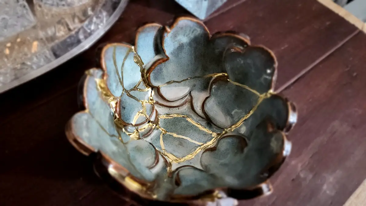 A close-up photo shows a ceramic bowl with a scalloped edge, featuring a blue-gray glaze and visible cracks repaired with…