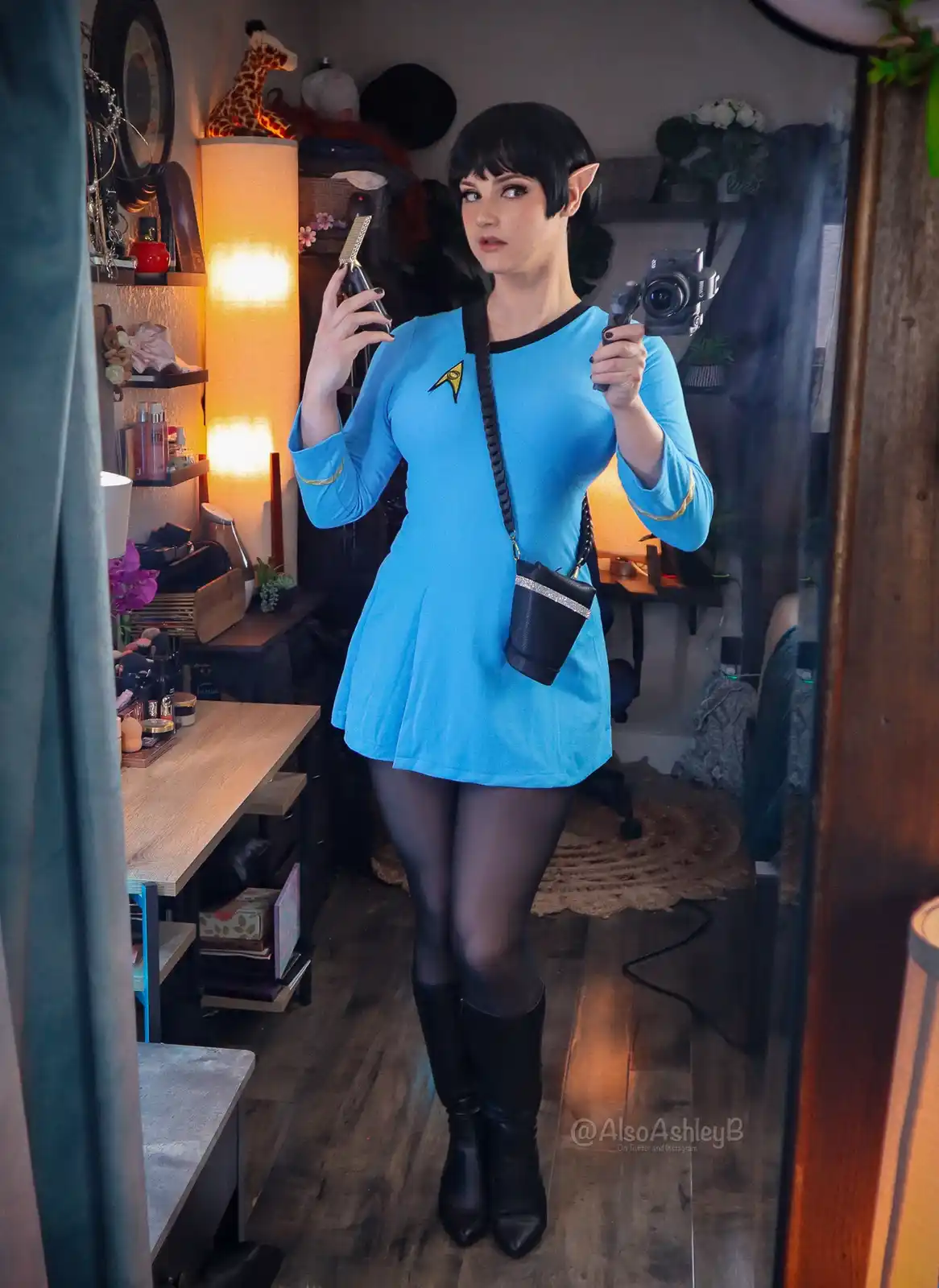 A person in a blue Star Trek uniform with pointed ears, holding a camera in one hand and a hair clipper in the other, looks…