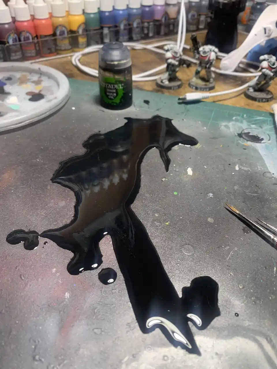 A spilled bottle of Citadel black paint on a grey crafting mat forms a shape resembling a dragon with wings, tail, and white…
