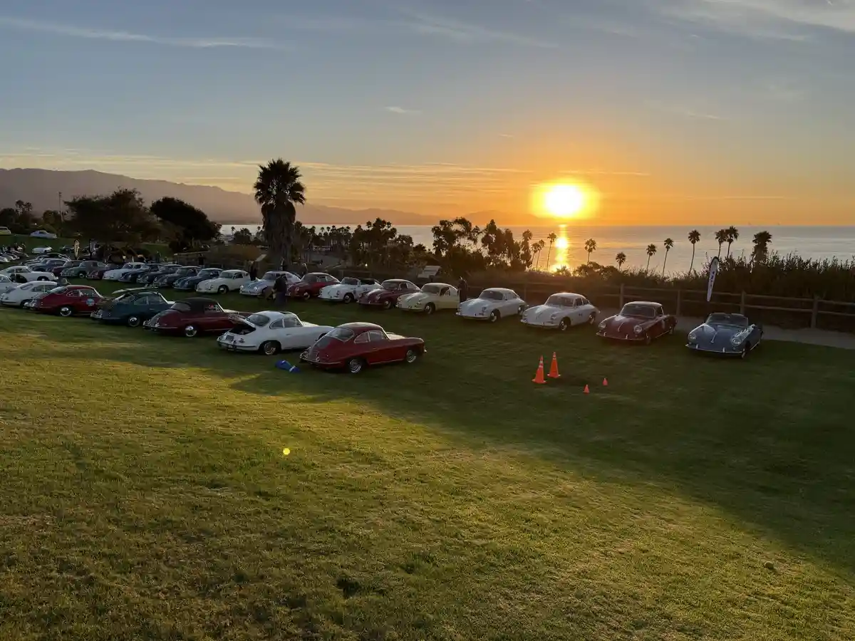 A scenic photo of numerous vintage Porsche cars parked on a grassy field overlooking the ocean at sunset, with palm trees…