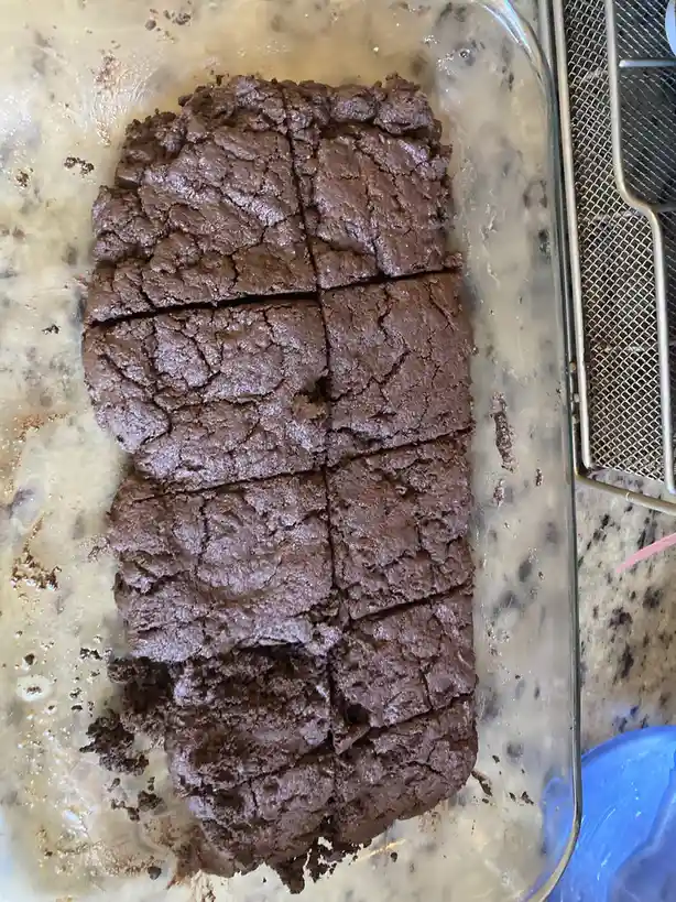 A close-up photo shows a glass baking dish containing freshly baked, dark chocolate brownies that have been cut into eight…