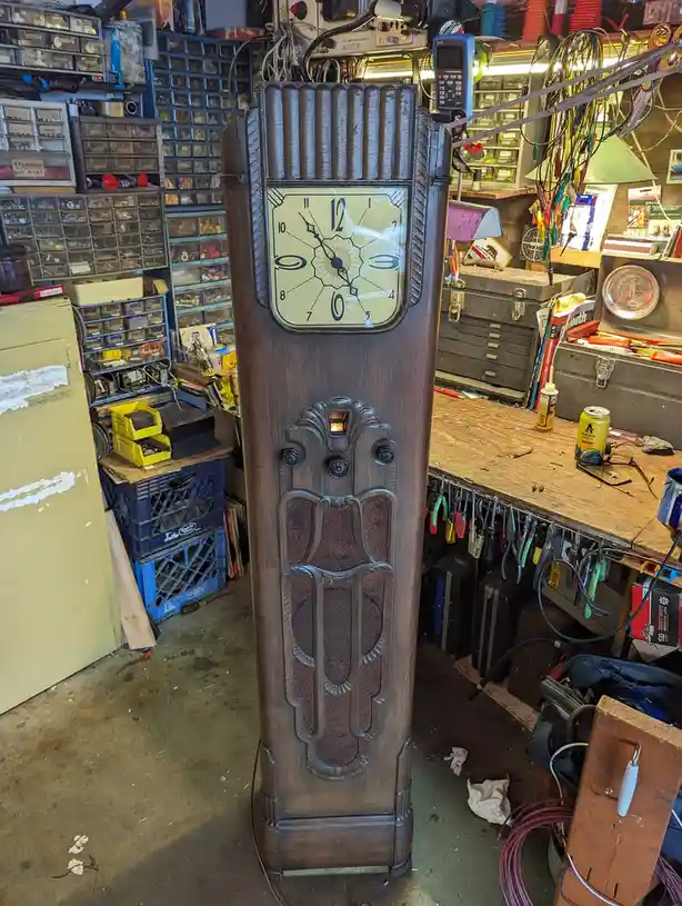 An antique wooden Art Deco clock stands in a cluttered electronics workshop filled with tools, parts, and equipment. The…