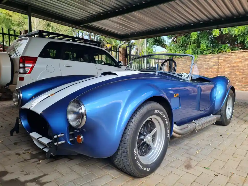 A shiny blue classic convertible sports car with white racing stripes is parked under a metal carport, with a white SUV…