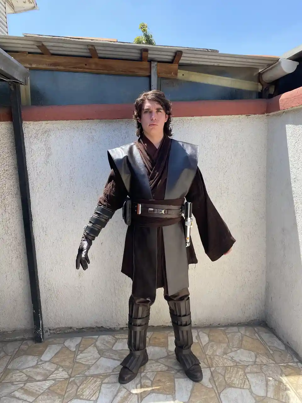 A young man in a detailed Anakin Skywalker cosplay from Star Wars stands outdoors against a plain wall, looking directly at…