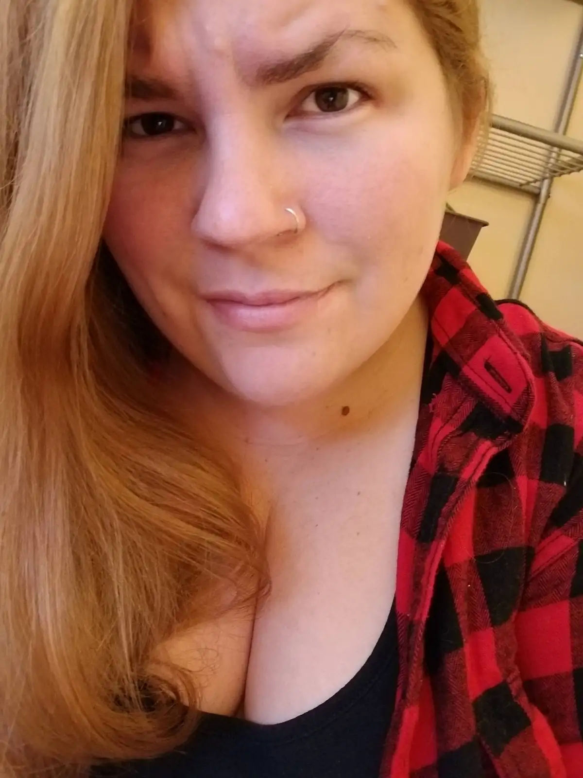 A close-up selfie of a woman with long blonde hair, wearing a red and black plaid shirt over a black top, looking directly…