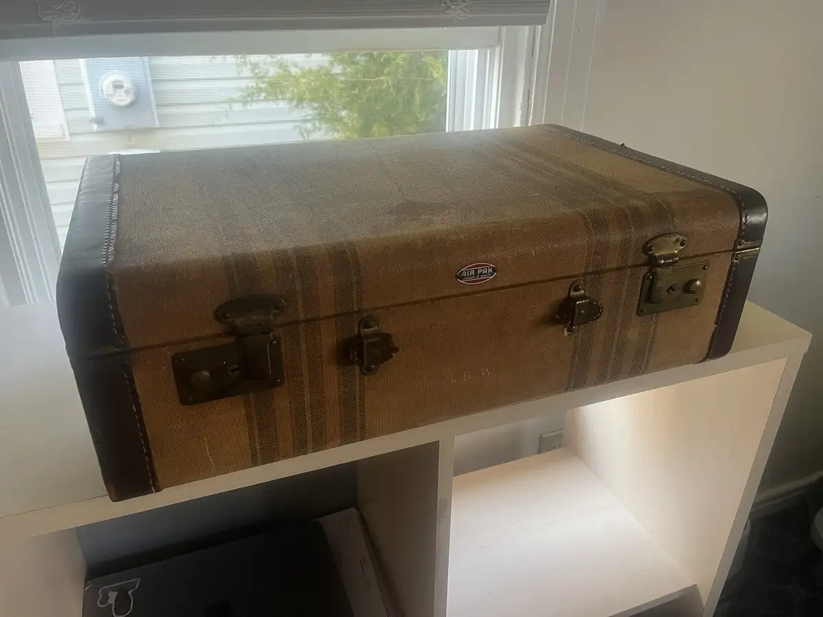 A vintage brown and tan striped suitcase with brass latches and an 'AIR PAK' logo sits on a white shelf, with a window and…