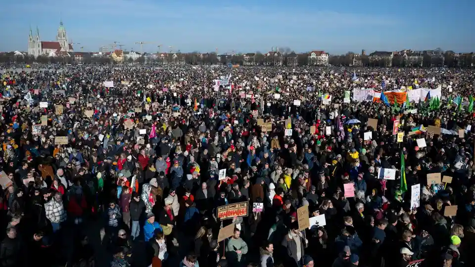A massive crowd of protesters fills a city square under a clear blue sky, holding numerous handmade signs and banners. In…