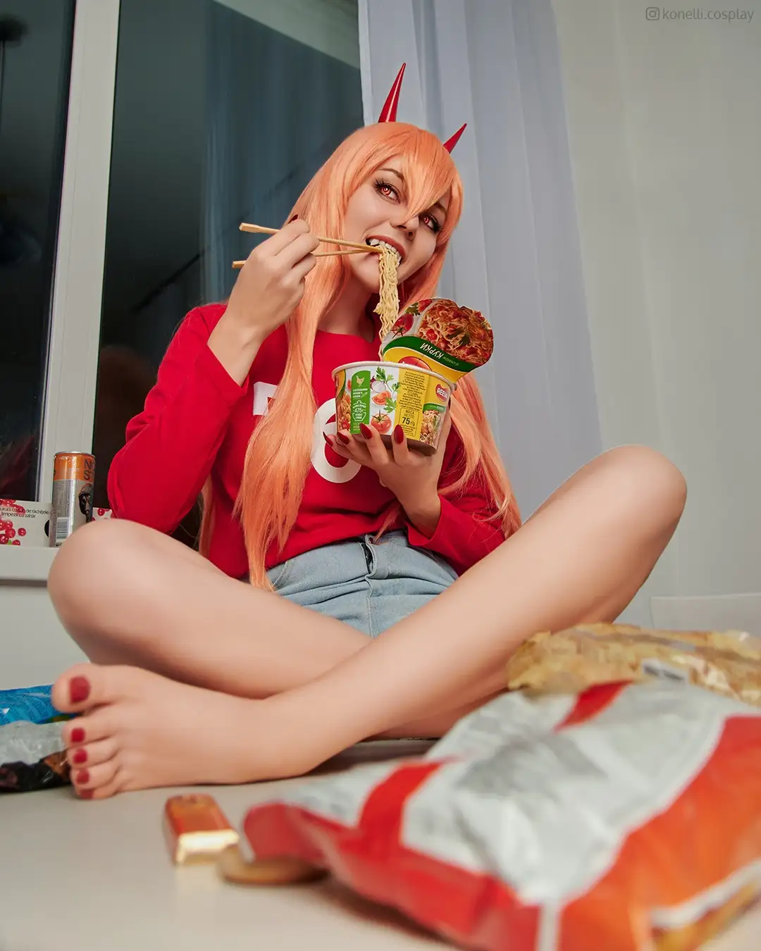 a person with long orange hair and red devil horns sits cross-legged on the floor eating ramen noodles with chopsticks…