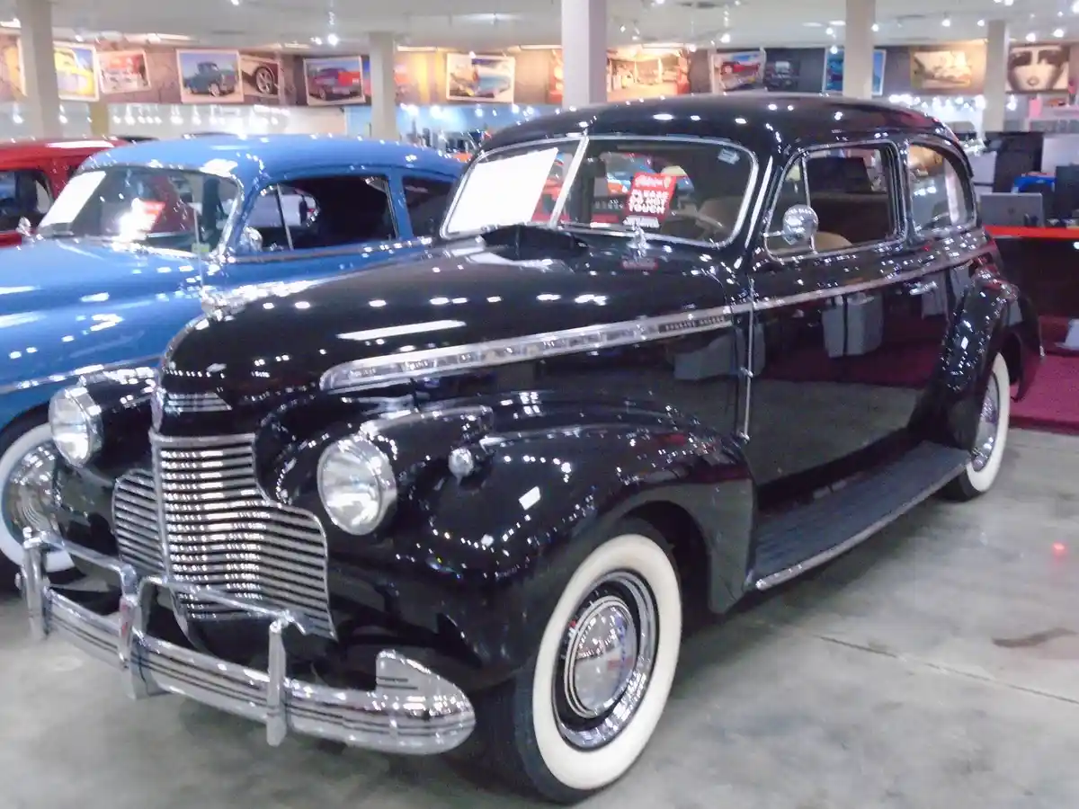 A glossy black vintage car with chrome accents and white-wall tires is displayed indoors, likely in a museum or showroom…