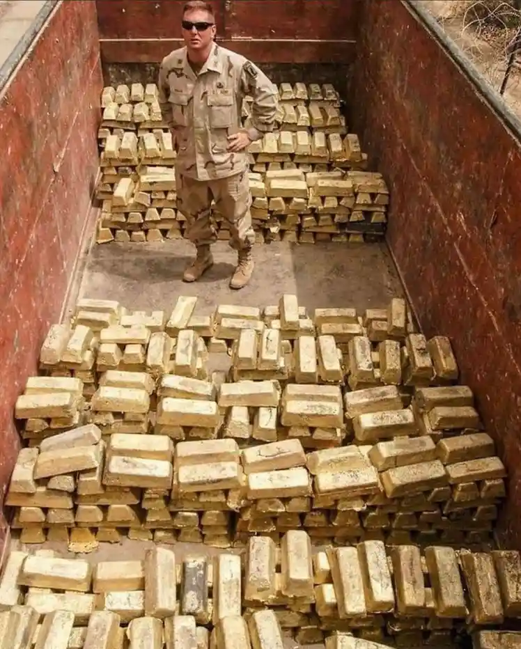 A man in a military uniform and sunglasses stands confidently with hands on hips inside a large truck bed filled with stacks…