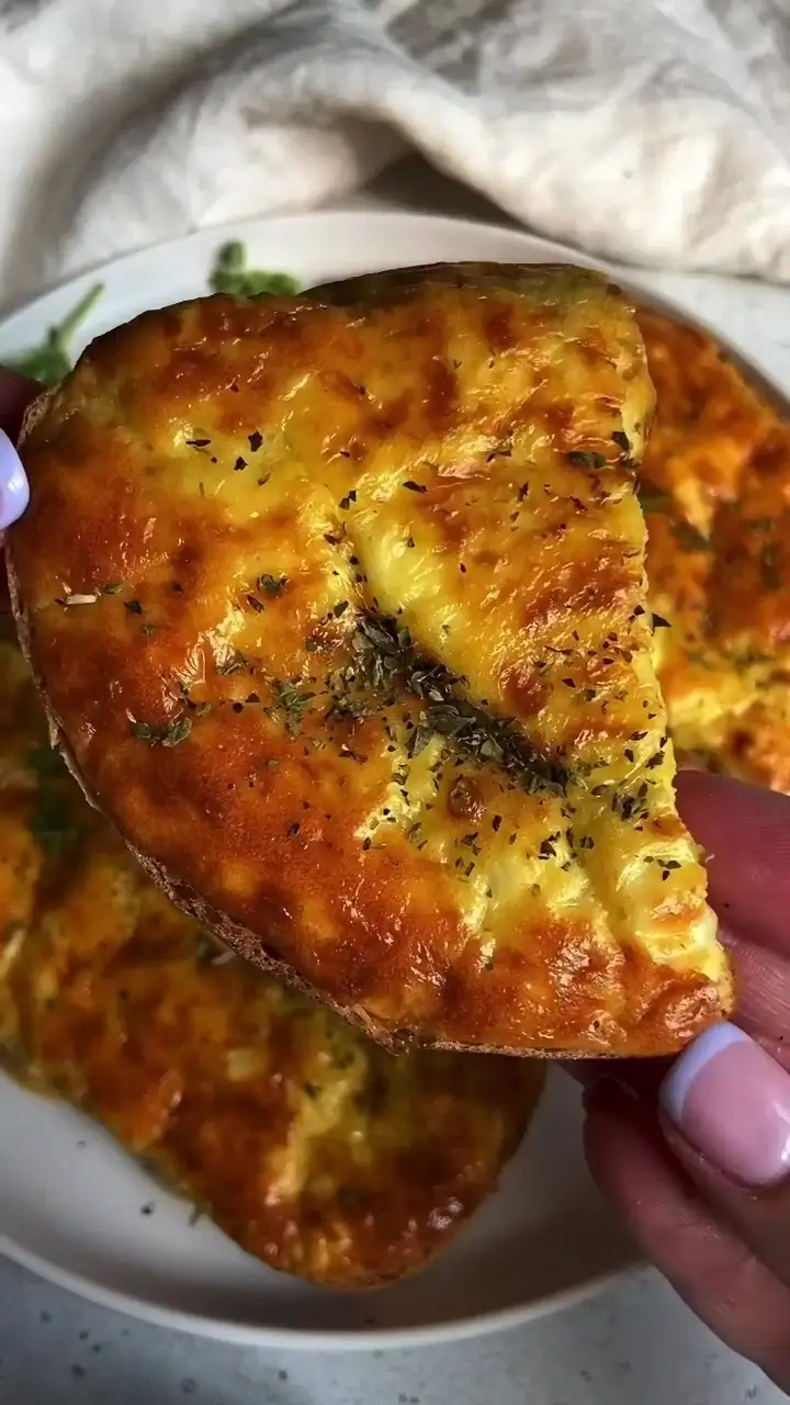 a close-up photo shows a hand with pink and white manicured nails holding a slice of golden-brown, cheesy baked bread or…