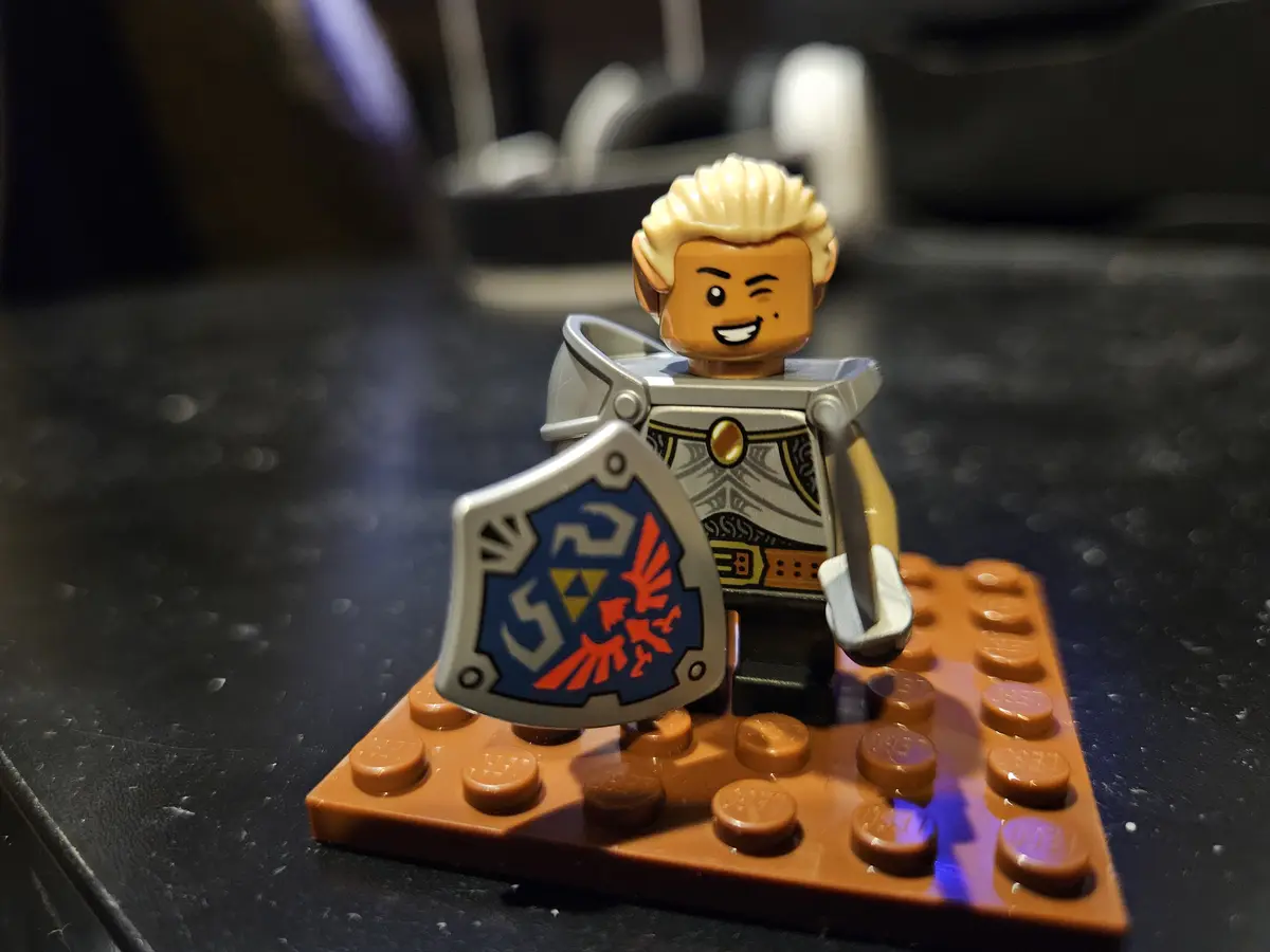 A close-up photo of a LEGO minifigure resembling Link from The Legend of Zelda, standing on a brown LEGO baseplate. The…