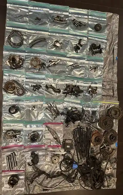 An overhead shot displays a large collection of disassembled engine parts, meticulously sorted into numerous labeled zip-top…