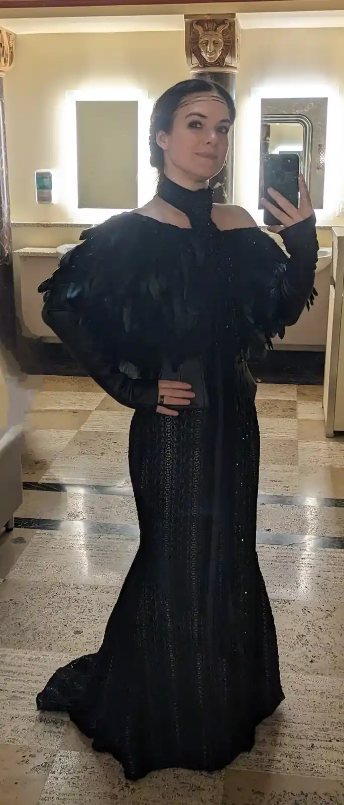 A woman in a dramatic black feathered gown takes a mirror selfie in a lavishly decorated bathroom with marble floors and…