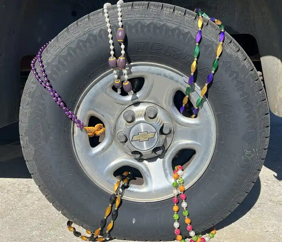 A close-up photo shows several strands of colorful Mardi Gras beads draped over a black tire with a silver Chevrolet wheel…