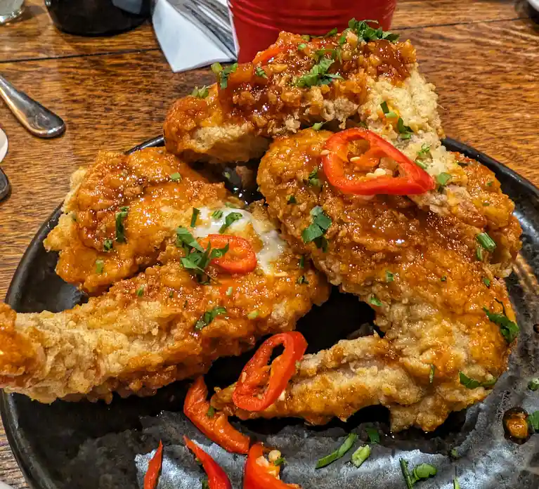 A close-up photo of a plate of crispy, golden-brown fried chicken pieces covered in a glossy orange sauce, garnished with…