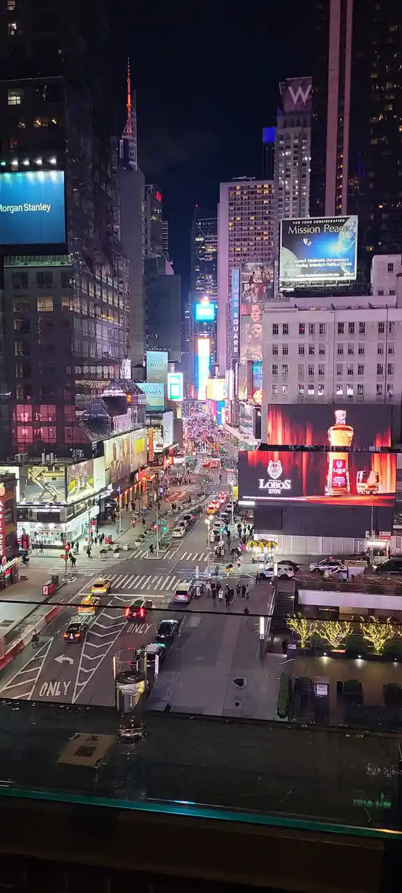A nighttime photo of Times Square, New York City, viewed from a high vantage point, with a glass of water on a glass ledge…