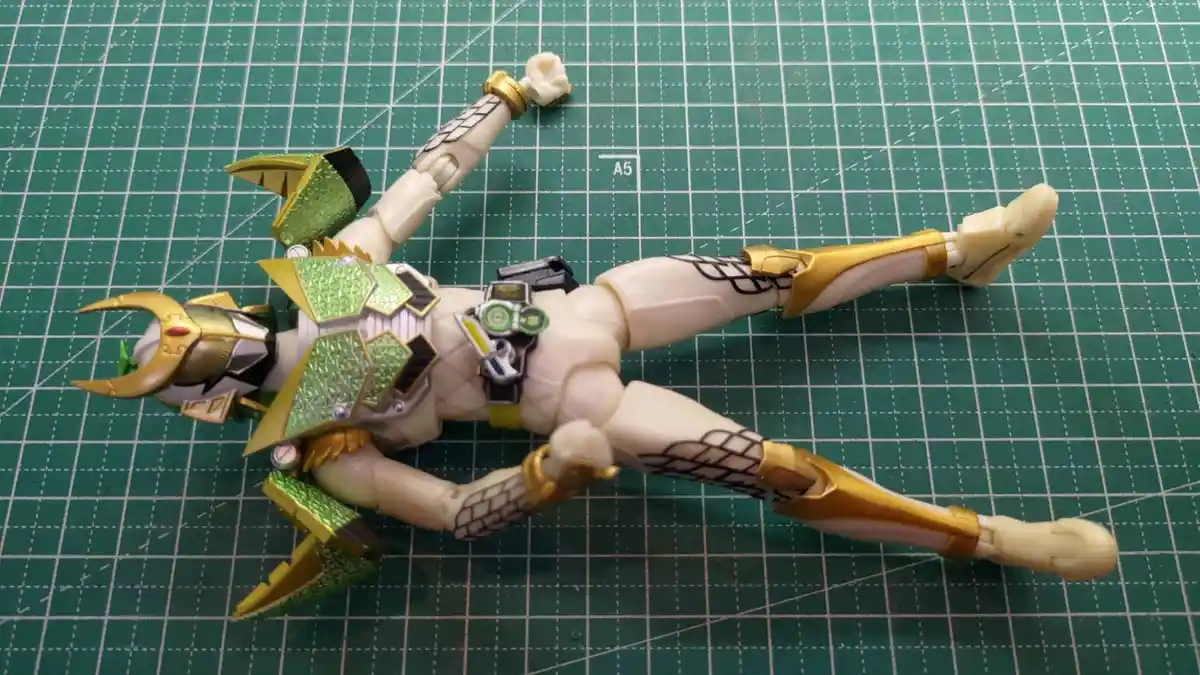 A green and gold armored action figure, resembling a Kamen Rider, lies flat on its back on a green grid cutting mat, limbs…